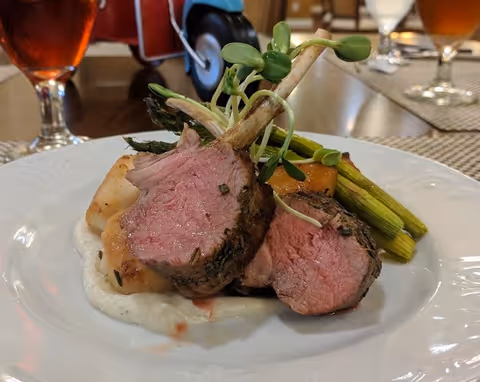 A plated gourmet meal featuring two pieces of herb-crusted lamb chops, served with grilled asparagus, a dollop of creamy white sauce, and garnished with microgreens on a white plate. In the background, there are glasses of iced tea and a dining table setting.