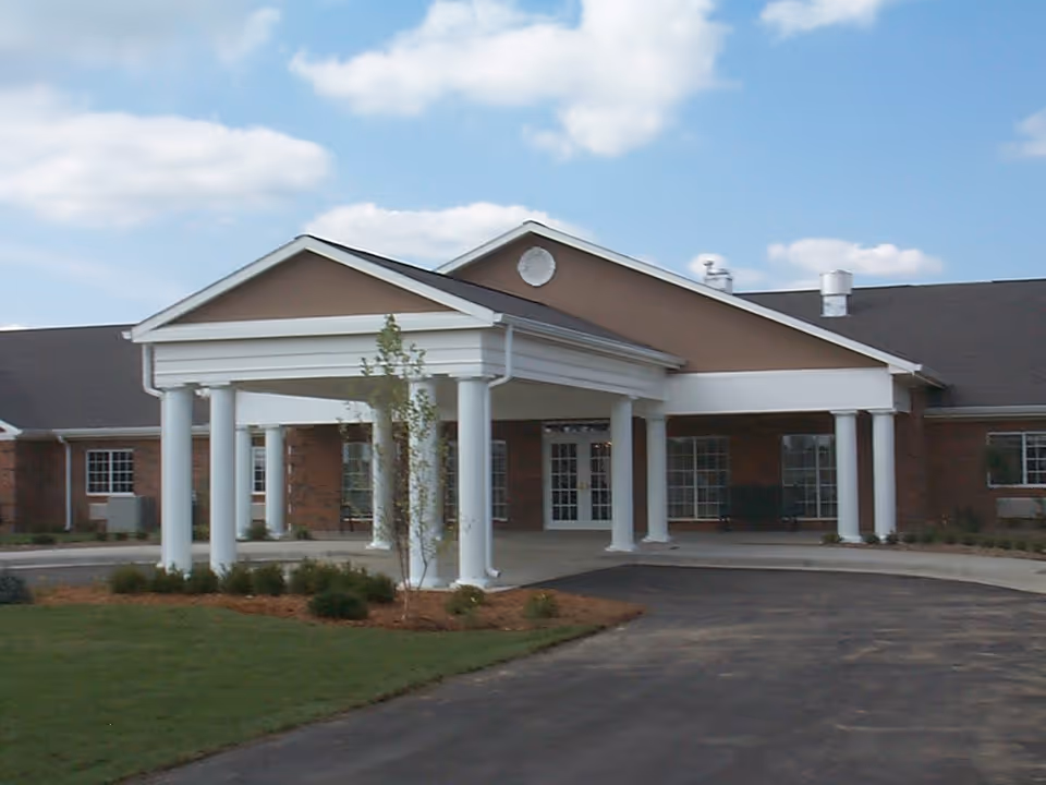 Front exterior view of Waterford Place Health Campus showing a covered entrance with white columns, brick walls, multiple windows, and a paved driveway under a partly cloudy sky.