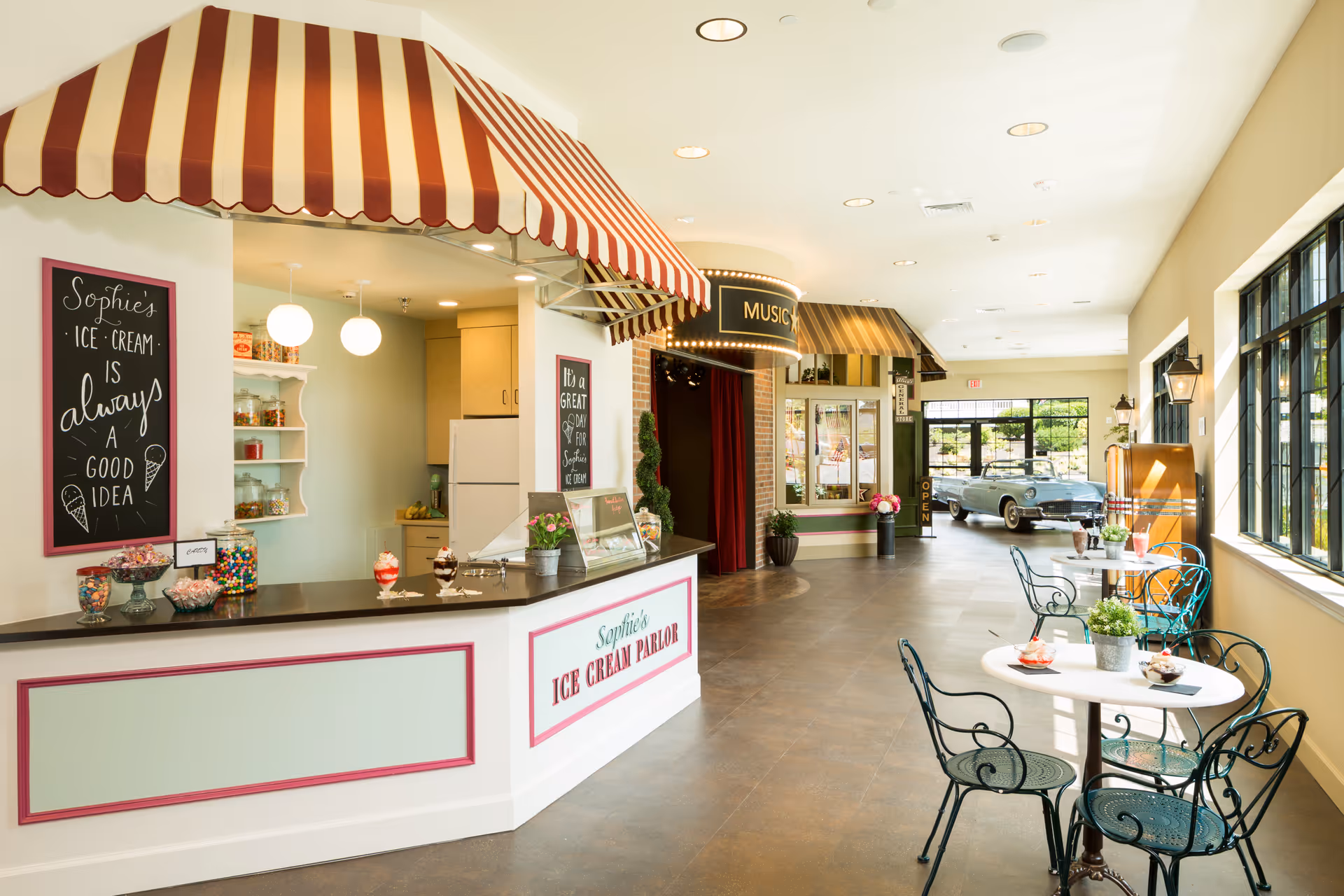 Interior view of an ice cream parlor area in a senior living facility with a counter displaying ice cream sundaes and candy jars. The counter has a red and white striped awning above it and signs that read 'Sophie's Ice Cream Parlor' and 'Sophie's Ice Cream is always a good idea.' There are small round tables with chairs and potted plants near large windows on the right side. Further down the hallway, there is a vintage car and a music-themed area with a marquee sign.