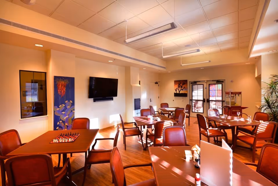 Bright communal dining and activity room with multiple tables and chairs, a wall-mounted TV, and a popcorn cart.