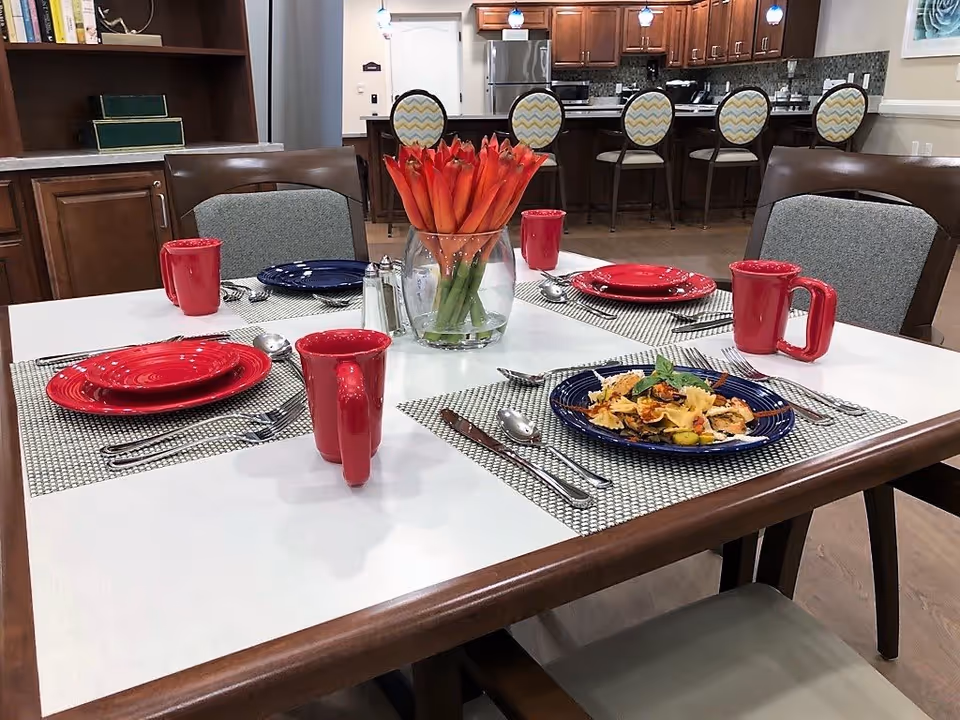 A dining table set for four with red mugs, red and blue plates, silverware, and a vase of orange flowers in the center. In the background, there is a kitchen area with wooden cabinets, a refrigerator, and a counter with four bar stools featuring patterned cushions.