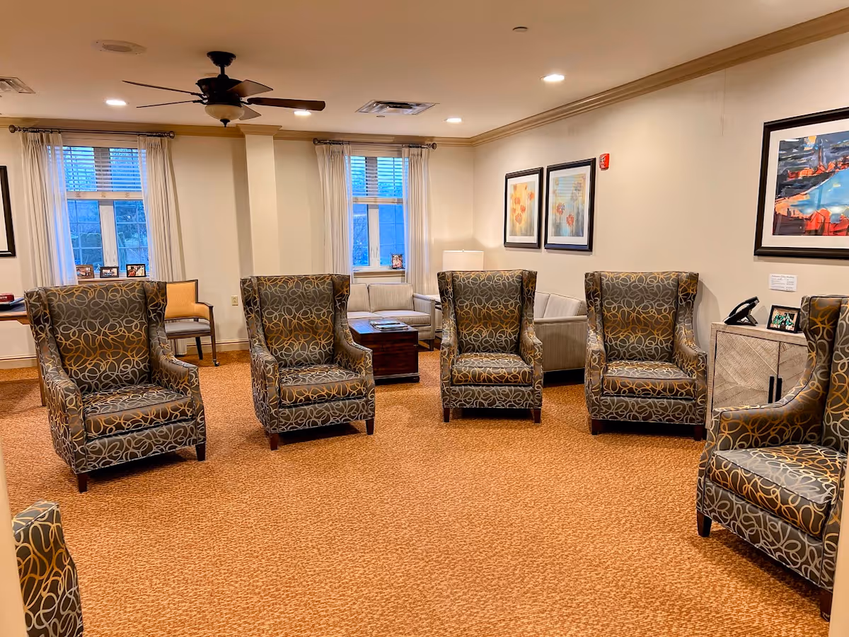 A cozy living room area with six patterned armchairs arranged in a semi-circle on a carpeted floor. The room has beige walls adorned with framed artwork, two windows with white curtains, a ceiling fan with lights, and a small table with a lamp and photo frames in the background.