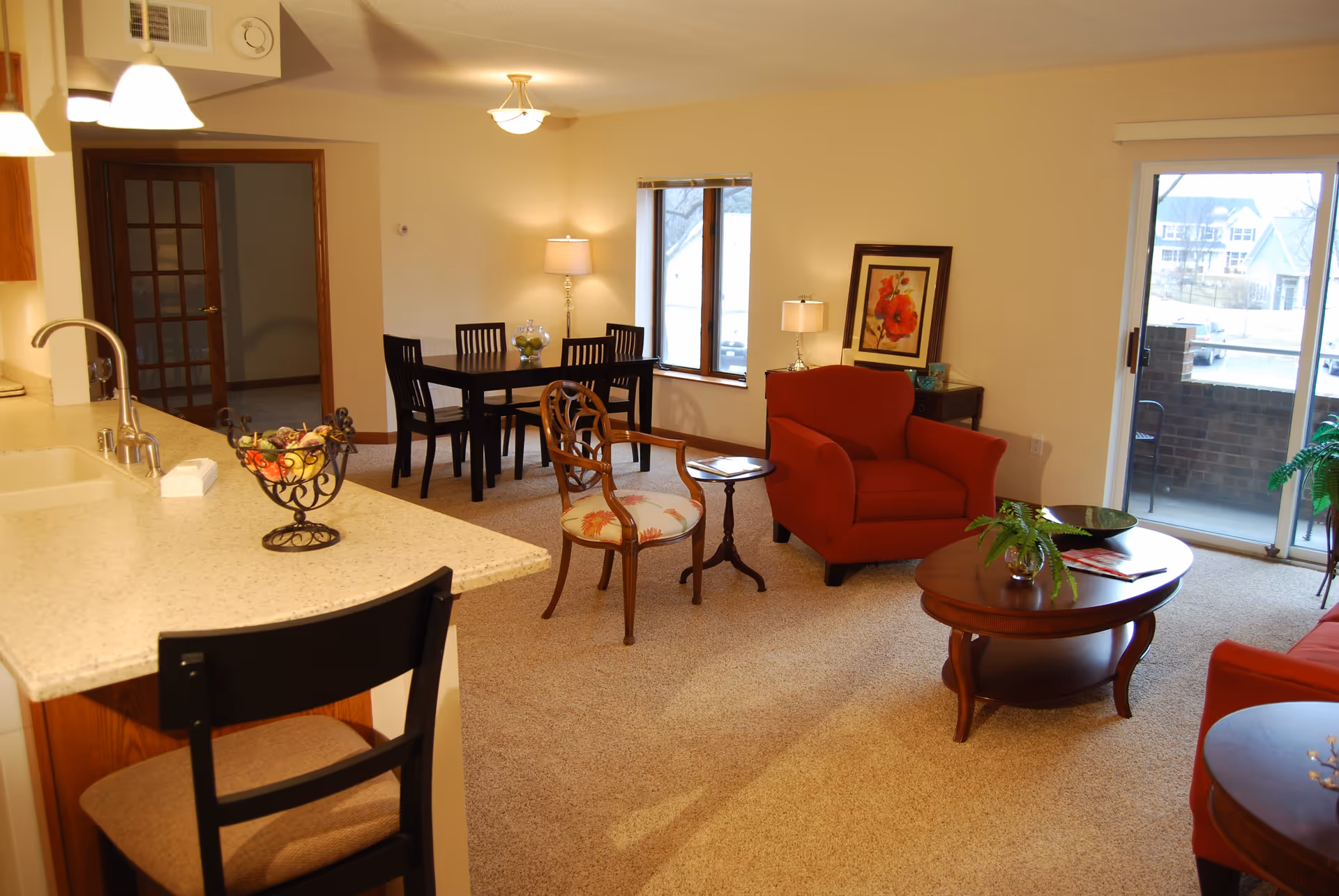 A cozy living and dining area in an assisted living facility. The room features a red armchair, a wooden coffee table with a small plant, a decorative chair with floral upholstery, and a small side table. In the background, there is a dining table with four chairs, a lamp, and a framed floral artwork on the wall. The kitchen counter with a sink and a bar stool is visible on the left side. Large windows and a sliding glass door provide natural light and a view of the outside.