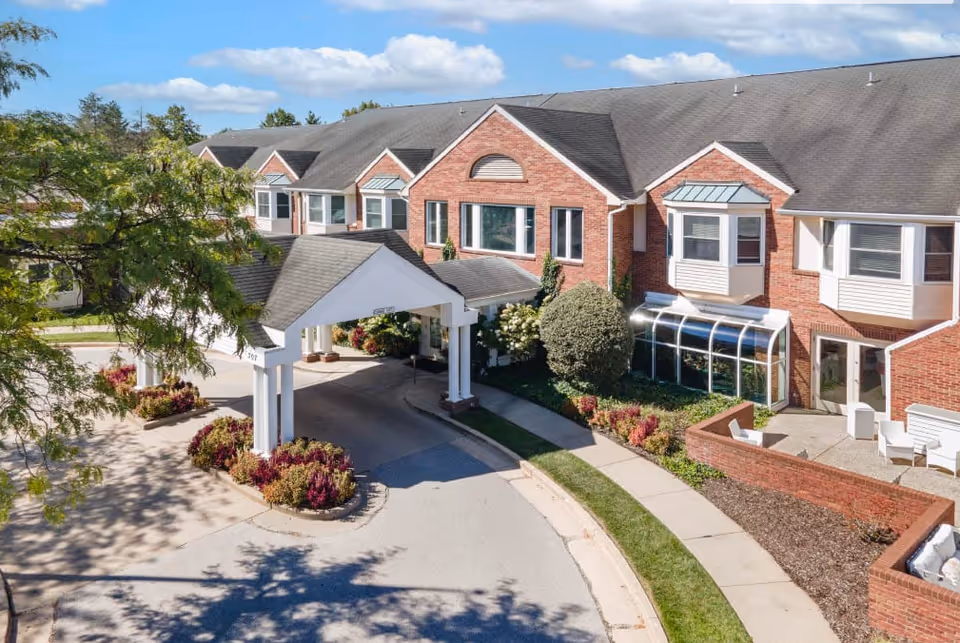 Exterior view of a senior living facility named Brooking Park featuring a brick building with multiple windows, a covered entrance with white pillars, landscaped bushes and flowers, a curved driveway, and a clear blue sky with some clouds.