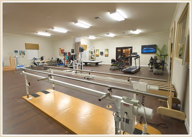 A spacious rehabilitation or physical therapy room with parallel bars for walking exercises in the foreground, various exercise equipment including treadmills, stationary bikes, and therapy tables in the background, bright overhead lighting, and framed pictures on the walls.