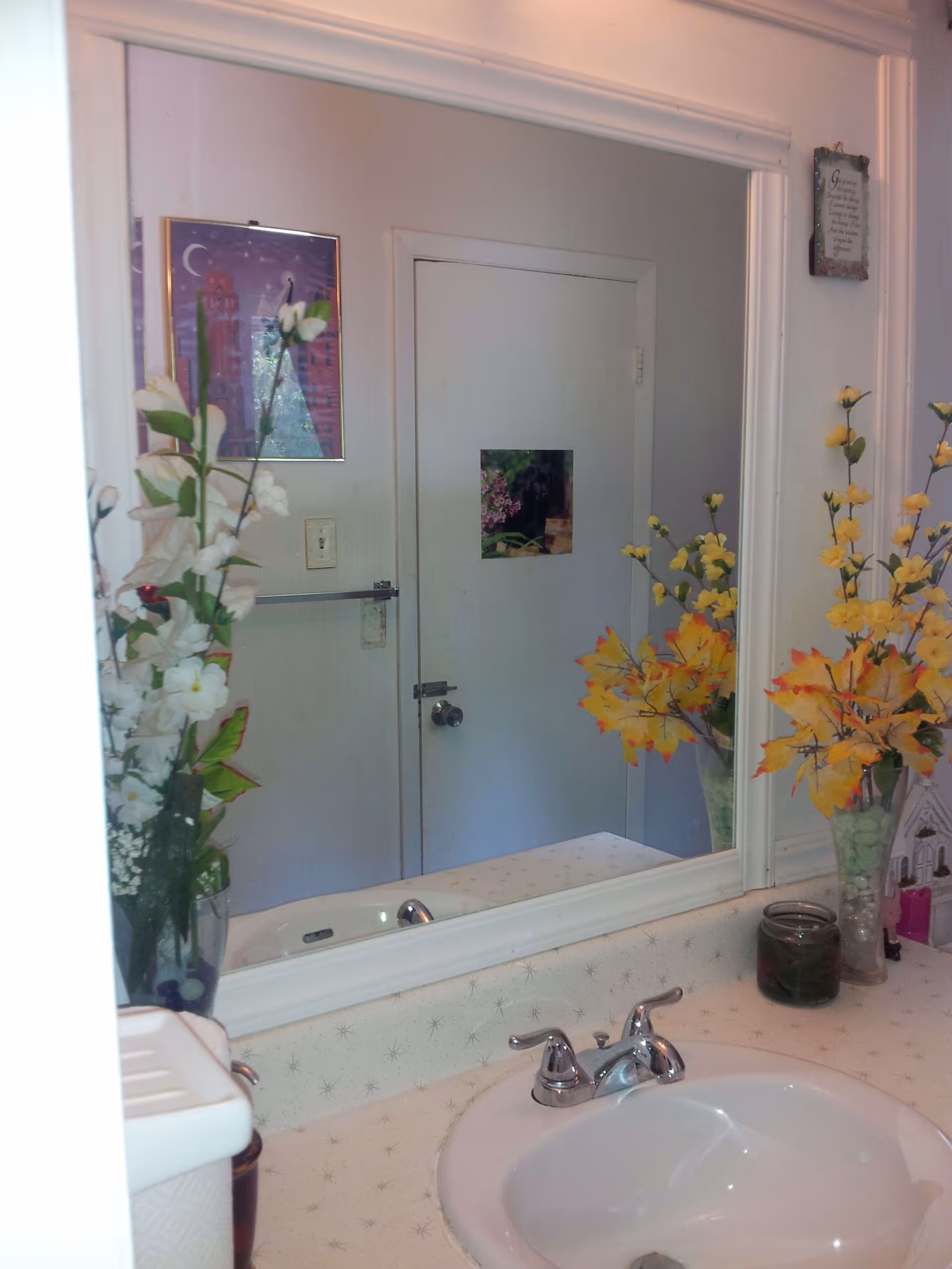 Bathroom sink area with a white countertop and silver faucet. A large mirror above the sink reflects a closed door with a small picture on it. On the countertop, there are two vases with artificial flowers, one with white flowers and the other with yellow and orange leaves. A small candle and a decorative item are also visible on the right side of the counter.