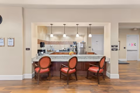 A clean and modern kitchen area with a granite countertop bar and three wooden chairs with red cushions. The kitchen has stainless steel appliances including a refrigerator and coffee machine, pendant lights hanging from the ceiling, and a tiled backsplash. The surrounding area has light-colored walls and wooden flooring.