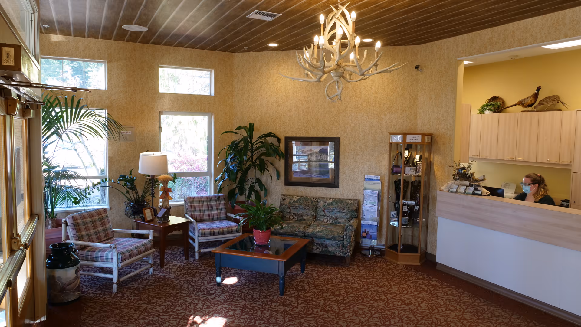 A cozy senior living facility lobby with patterned carpet, two plaid armchairs, a floral sofa, a wooden coffee table with a potted plant, and several large green plants near windows. A receptionist wearing a mask is seated behind a light wood reception desk. The walls are beige with a framed picture and a glass display cabinet. An antler chandelier hangs from the wooden ceiling.