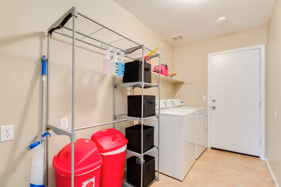 Small laundry room with a washer and dryer, metal shelving, cleaning supplies, and two red bins.