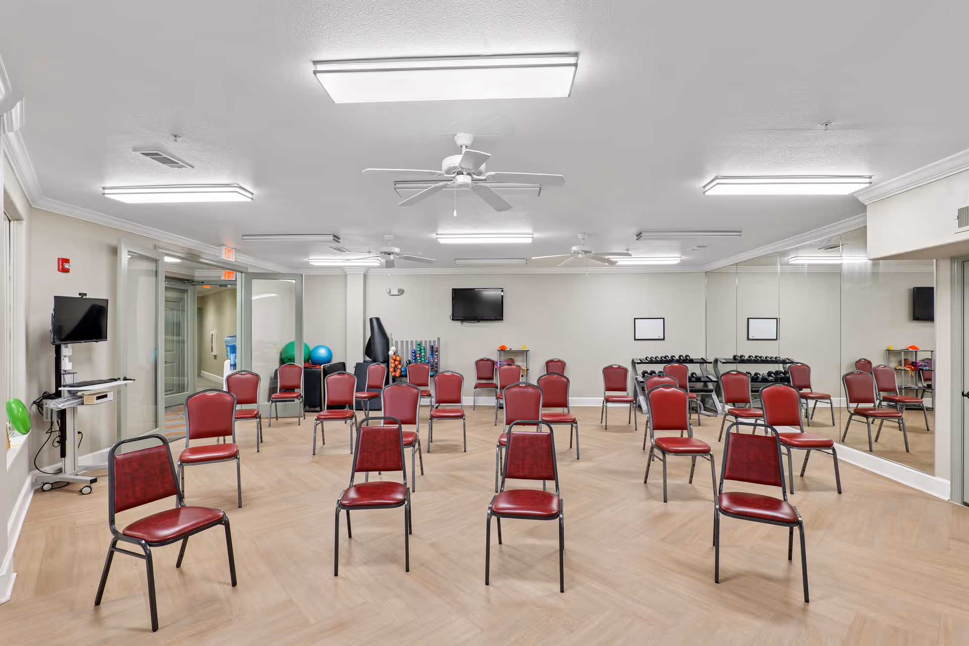 A spacious room with multiple red chairs arranged in rows facing a wall-mounted TV. The room has light wood flooring, white walls, and a large mirror covering one wall. Exercise equipment such as dumbbells and exercise balls are visible in the back corner. Ceiling fans and fluorescent lights are mounted on the ceiling.