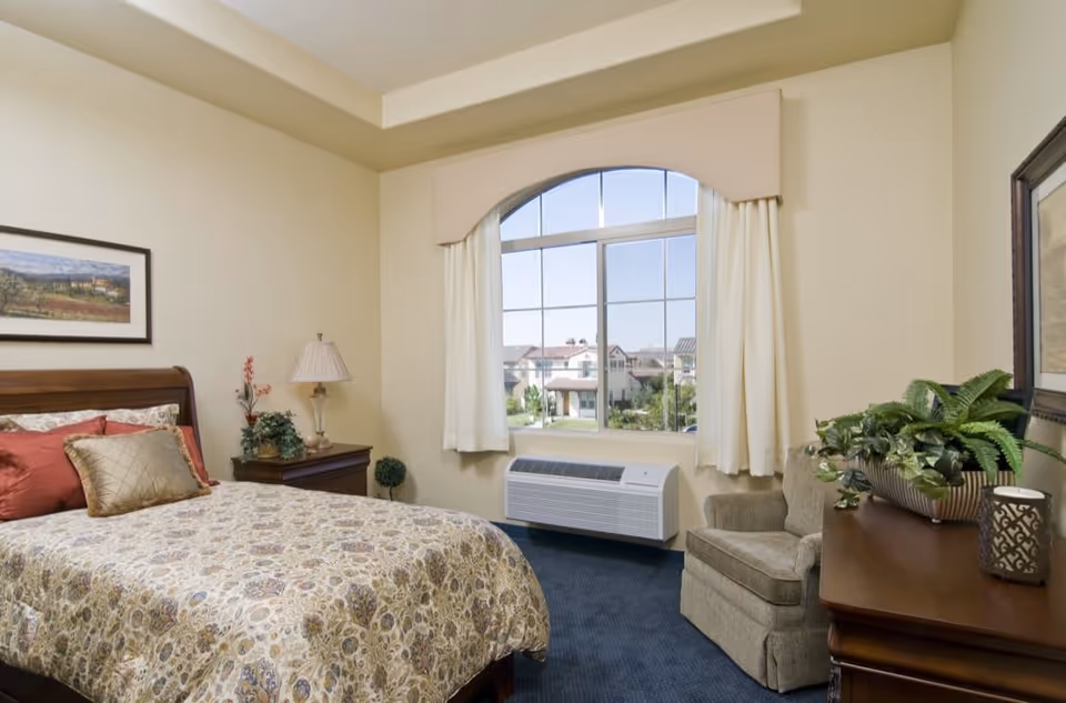 Neatly furnished bedroom with a made bed, nightstand and lamp, armchair, large arched window overlooking nearby houses, and a dresser with plants.