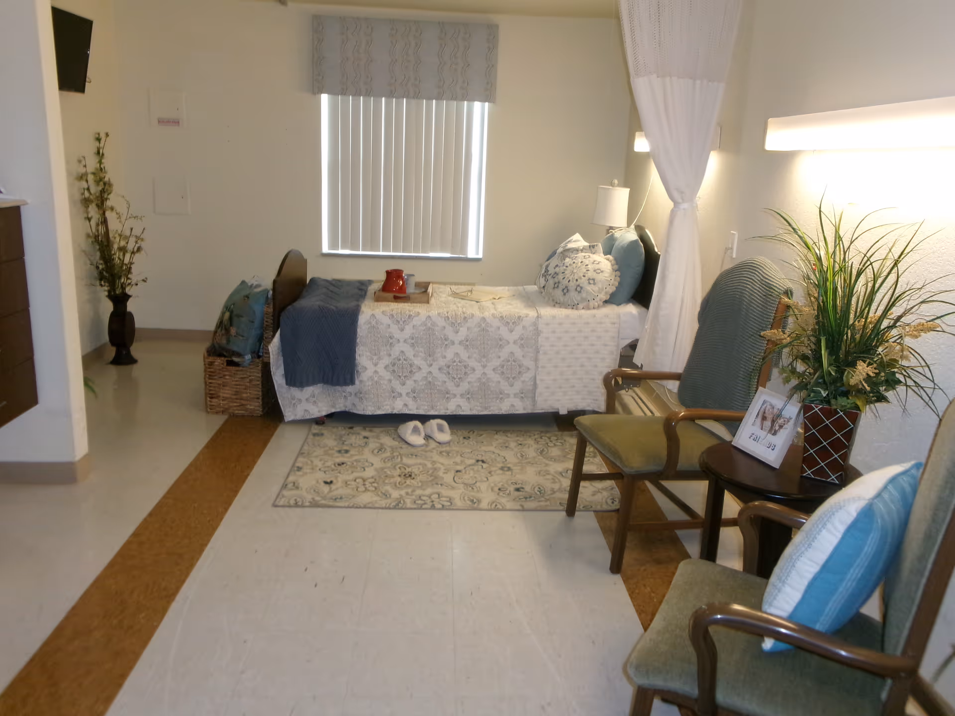 Small furnished patient room with a single bed, two chairs, a side table with a plant and a rug on the floor.