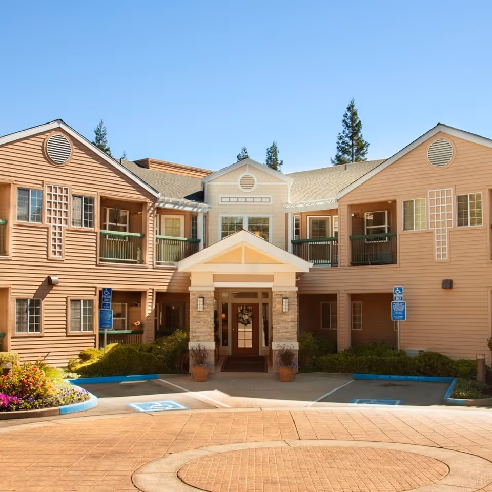 Front entrance of a two-story senior living building with a covered portico, balconies, landscaping, and accessible parking spaces.