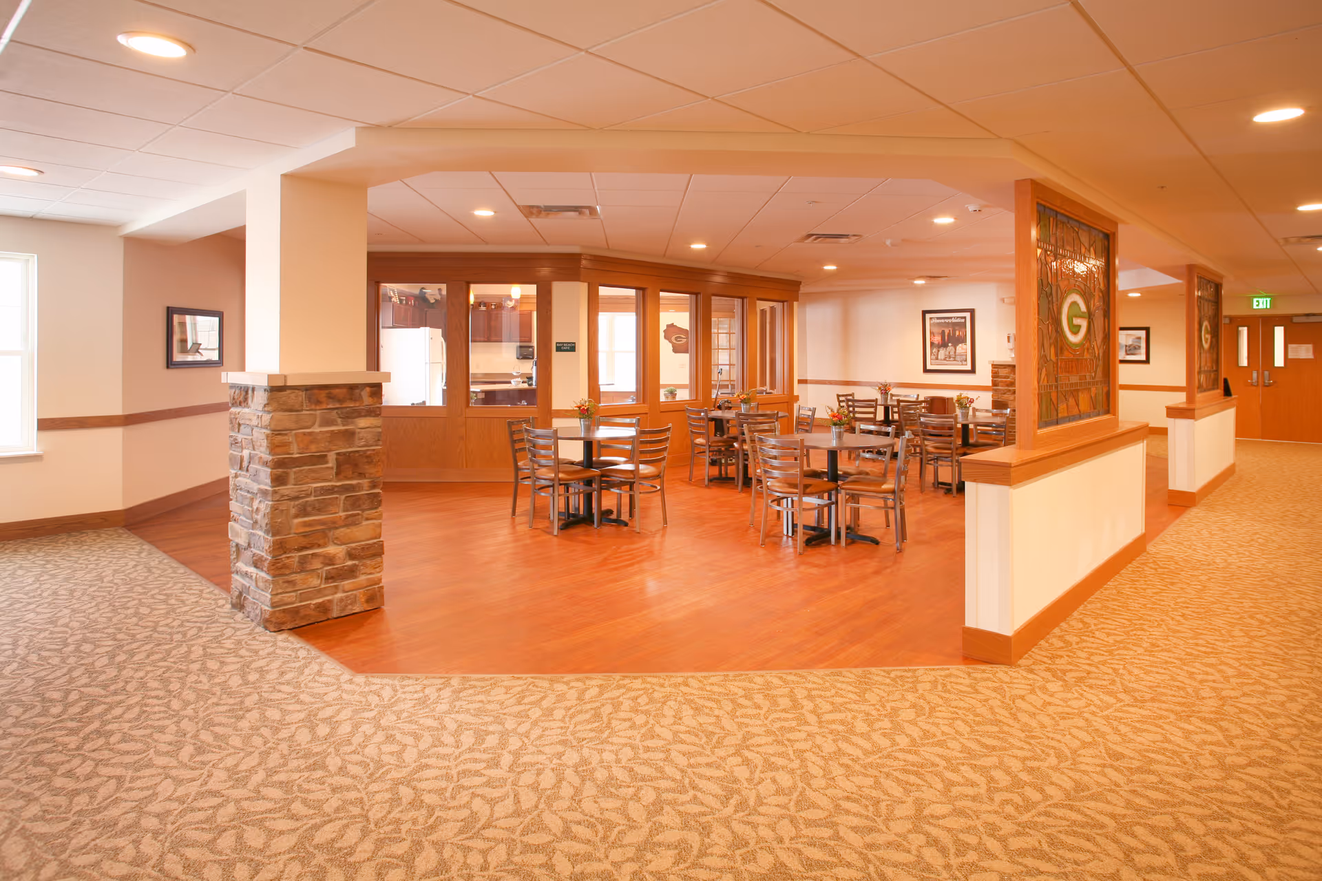 Open dining area with multiple round wooden tables and metal chairs, stained-glass panels, and warm wood flooring in a senior living common space.