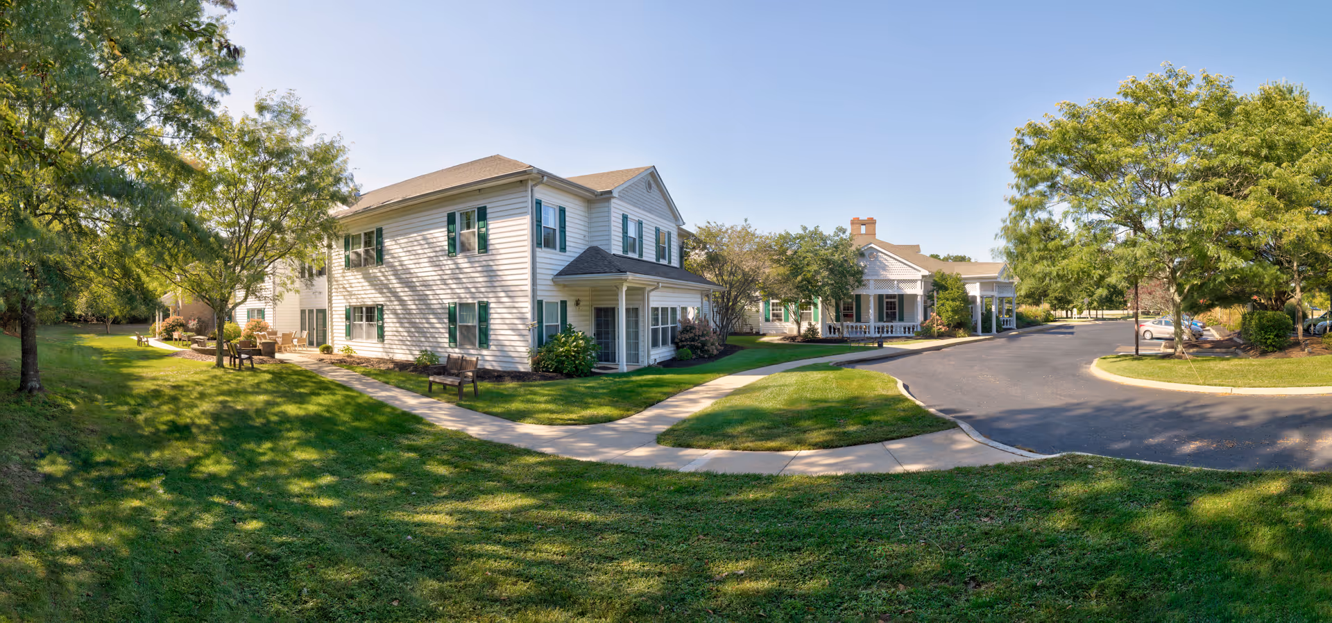 Exterior view of The Residence at Stafford showing white two-story buildings with green shutters, walkways, a paved driveway, lawn and trees under a clear sky.