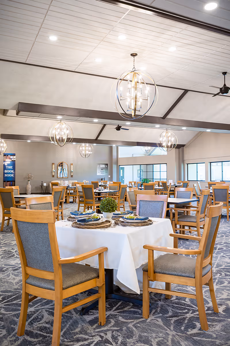 A spacious dining room in a senior living facility with multiple wooden tables and chairs. One table in the foreground is set with a white tablecloth, woven placemats, blue napkins, and a small green plant centerpiece. Modern spherical chandeliers hang from the ceiling, and large windows allow natural light to fill the room.