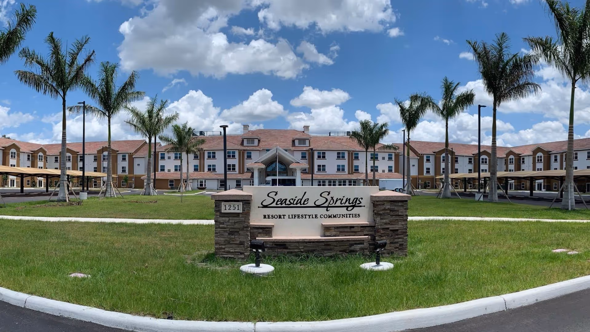 Front exterior of Seaside Springs Retirement Community showing the building, entrance sign on a grassy lawn, and palm trees under a blue sky.
