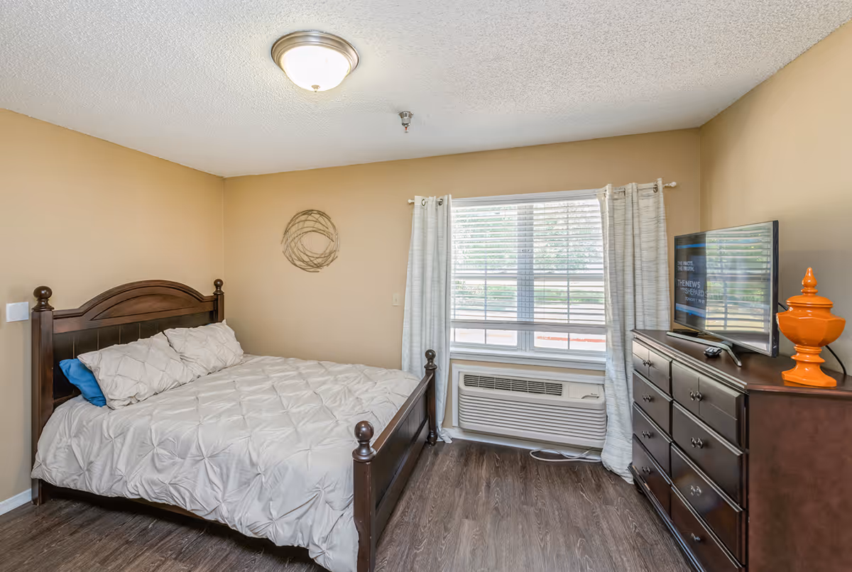 A bedroom with a wooden bed frame and a white comforter, a window with white curtains and blinds, a dark wooden dresser with a flat-screen TV and an orange decorative item on top, and wood flooring.