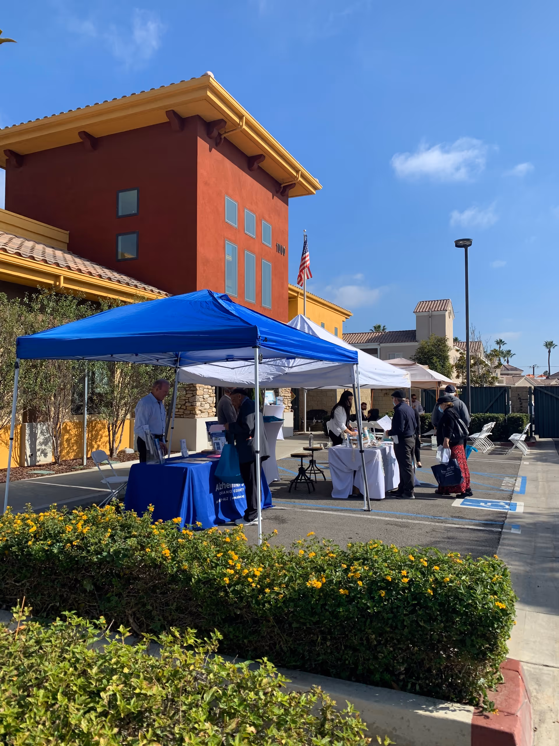 Outdoor scene showing a senior living facility with a red and yellow building in the background. Several people are gathered under blue and white canopy tents set up in a parking lot, with tables displaying informational materials. Yellow flowering bushes are in the foreground, and the sky is clear with a few clouds.