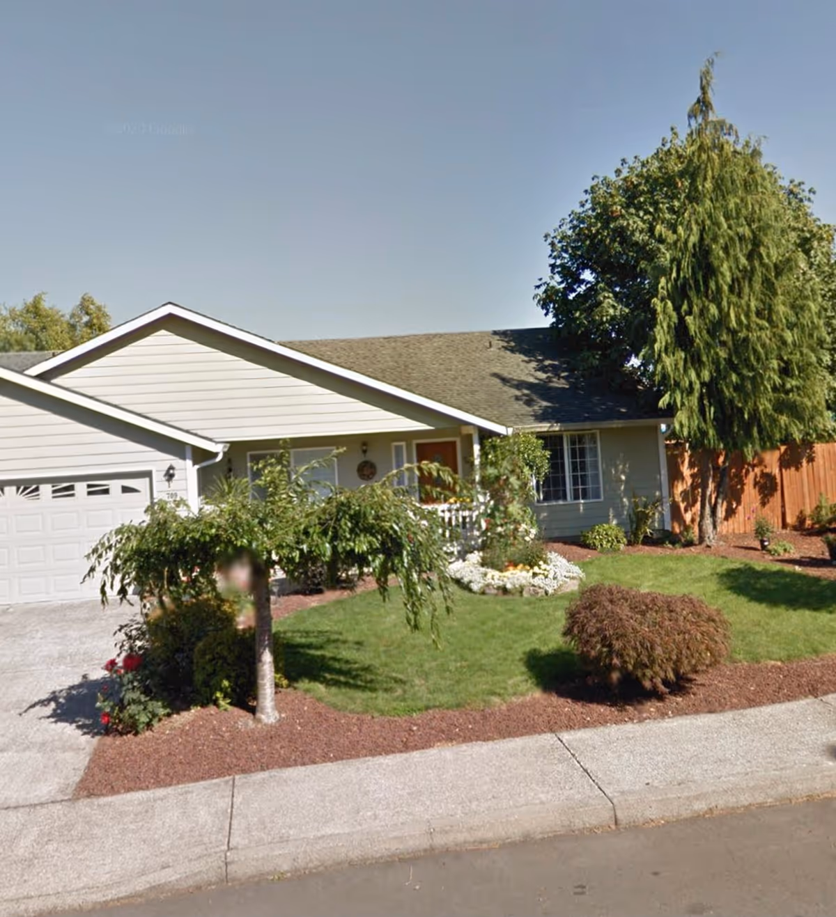 Front exterior of a single-story house with a garage, lawn, trees, and a sidewalk.