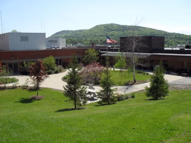 Brick building with a landscaped courtyard and picnic table, an American flag, grassy lawn and hills in the background.