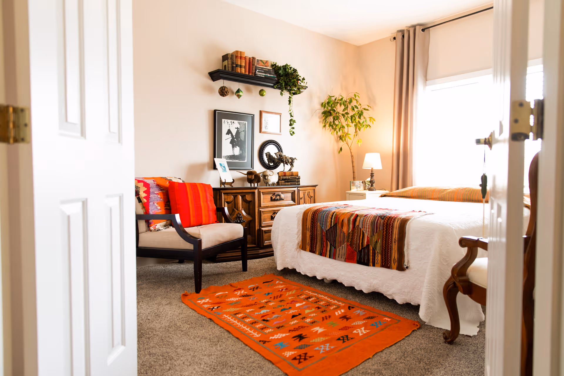 A cozy bedroom with a bed covered in a white quilt and a colorful patterned throw blanket. There is a wooden dresser with decorative items and framed pictures on the wall above it. A comfortable armchair with a red cushion and a patterned throw is next to the dresser. A small table lamp and a potted plant are near the window with beige curtains. An orange patterned rug is on the carpeted floor.
