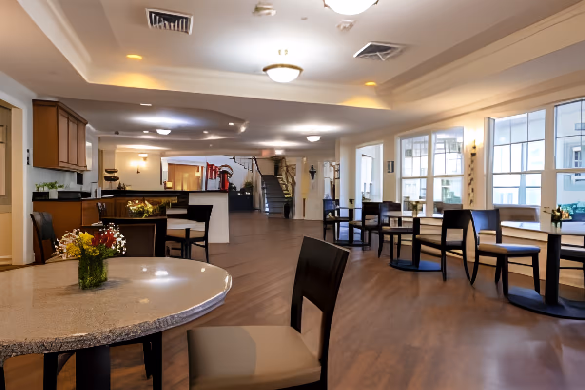 A spacious and well-lit common area with multiple tables and chairs arranged for seating. The room features large windows allowing natural light to fill the space, a countertop with cabinets on the left side, and a staircase in the background. Small flower arrangements are placed on the tables, creating a welcoming atmosphere.