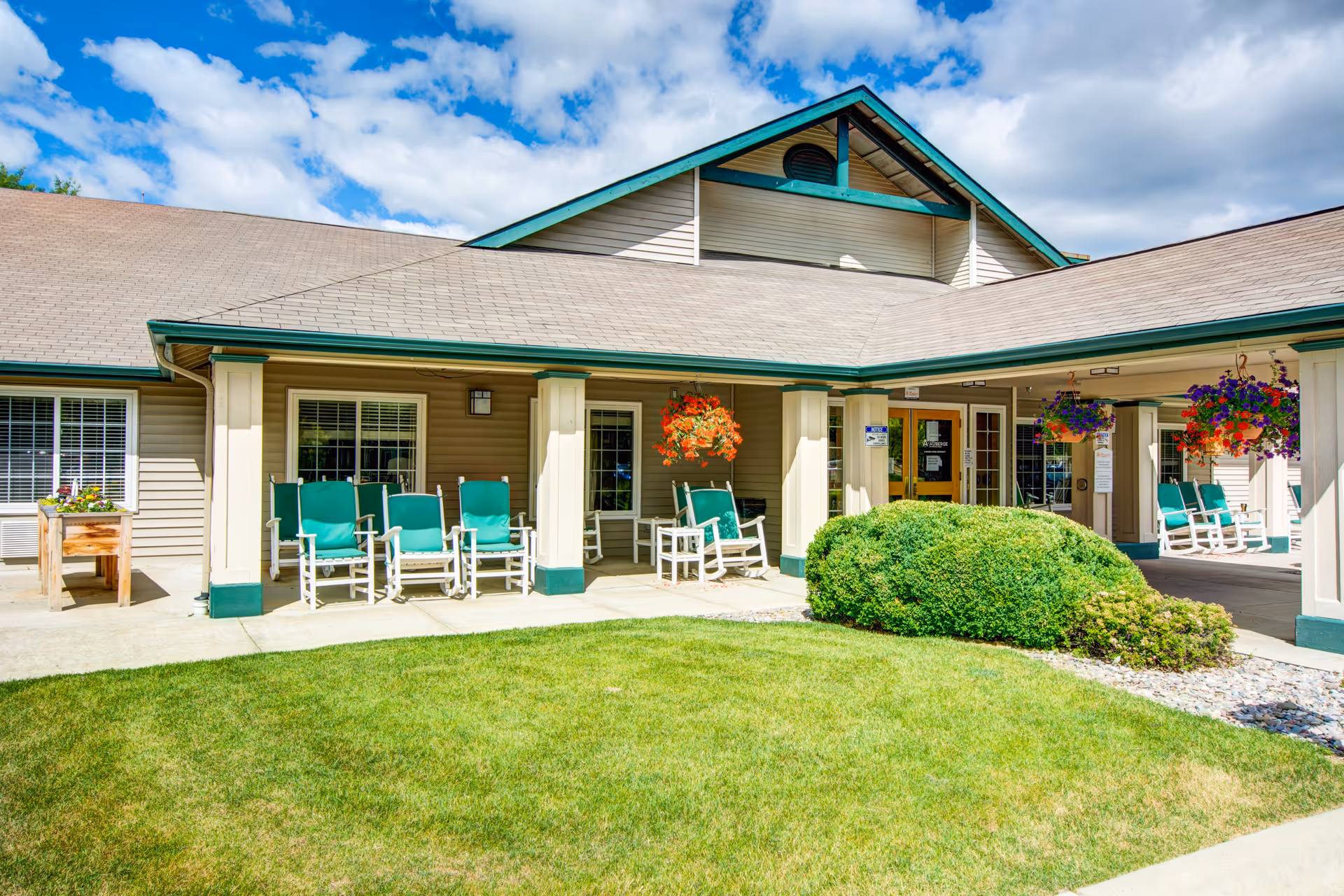 Exterior view of Missoula Valley Senior & Assisted Living facility showing a covered porch area with green cushioned rocking chairs, hanging flower baskets, and well-maintained landscaping including green grass and bushes under a partly cloudy blue sky.