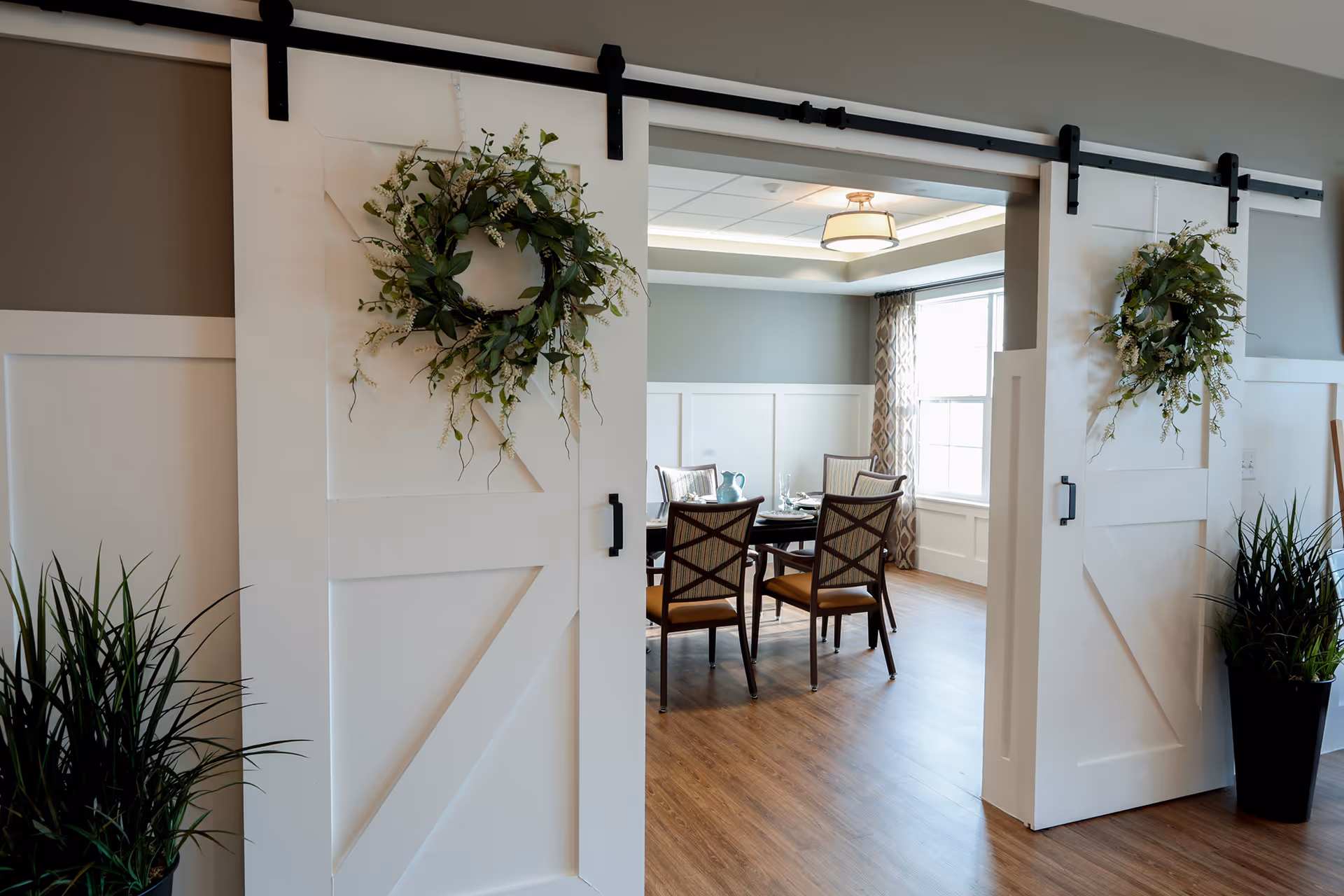 White sliding barn doors with wreaths open onto a dining room with a table and chairs.