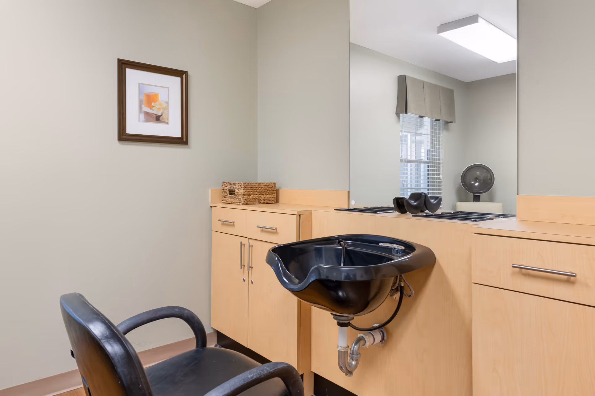 Interior of a hair washing station with a black salon chair and a black sink basin attached to a light wood cabinet. A large mirror is mounted on the wall above the cabinet, reflecting a window with blinds and a small fan. A framed picture hangs on the adjacent wall.