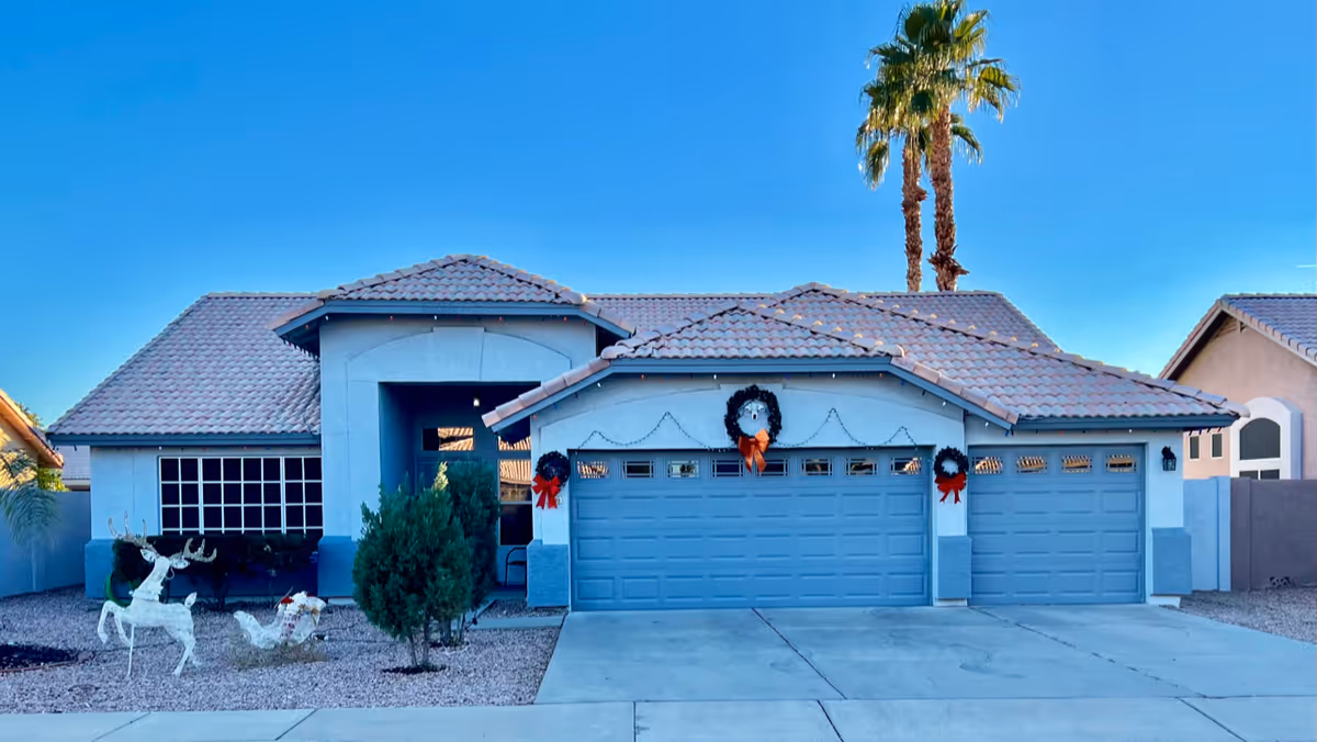Front exterior view of a single-story house with a tiled roof, three-car garage decorated with holiday wreaths and red bows, a small tree and holiday decorations in the gravel yard, and two tall palm trees in the background under a clear blue sky.