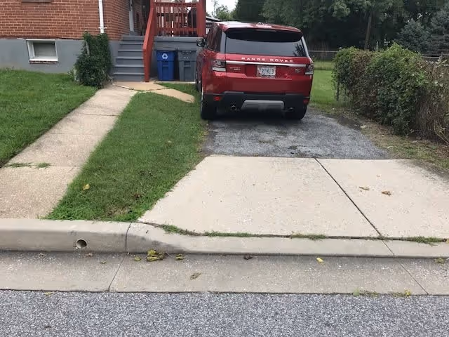 A red Range Rover parked on a narrow driveway next to a sidewalk and a small grassy lawn leading to a brick building with stairs and a porch. There are two blue recycling bins near the stairs and a hedge on the right side of the driveway.