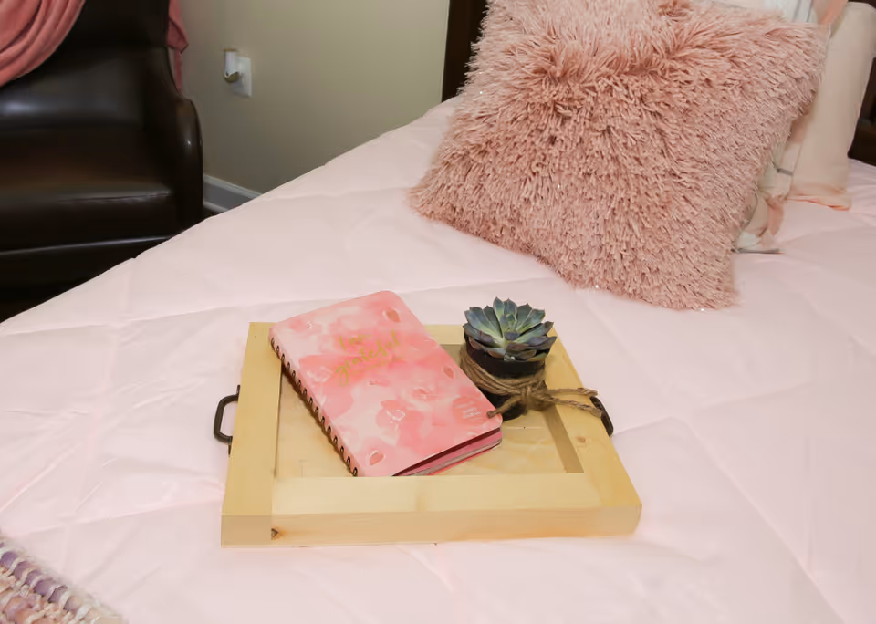 A close-up view of a bed with a light pink quilted bedspread, a fluffy pink pillow, and a wooden tray placed on the bed. On the tray, there is a pink floral notebook and a small potted succulent plant wrapped with twine. A dark brown leather chair and a pink blanket are partially visible in the background.