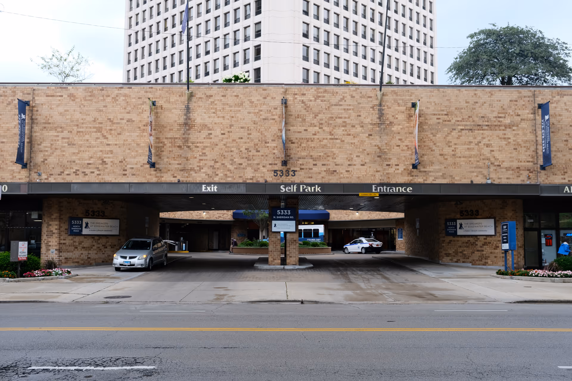 Exterior view of The Breakers at Edgewater Beach senior living facility showing a brick building with a covered driveway area for self parking, entrance, and exit. Two cars are parked under the covered area, and signs display the address 5333 N. Sheridan Rd.