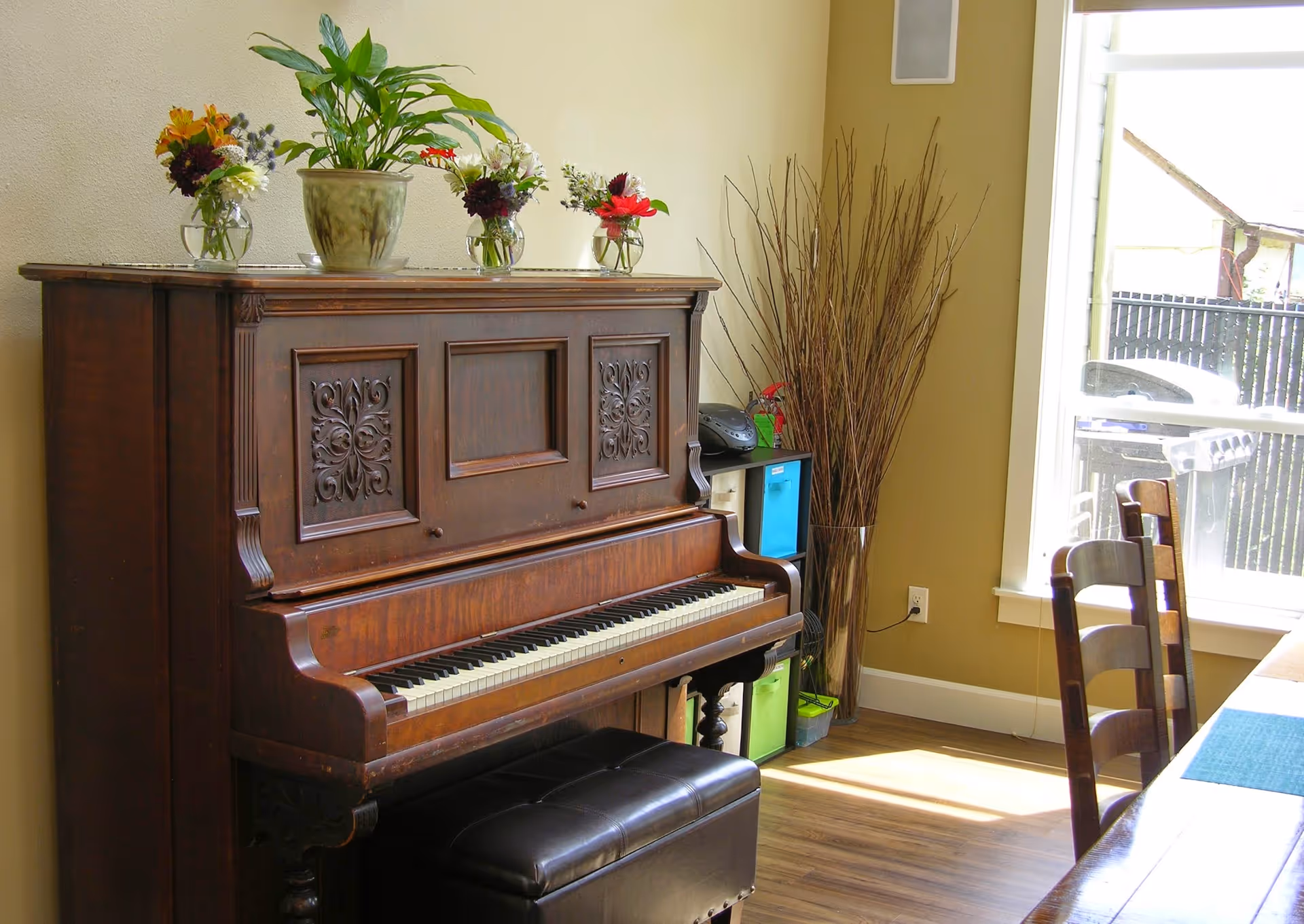 A wooden upright piano with intricate carvings on the front panel, topped with several small flower arrangements and a potted plant. Next to the piano is a small shelving unit with colorful storage bins and a tall vase filled with decorative branches. Sunlight streams in through a large window, illuminating wooden chairs and part of a table in the foreground.