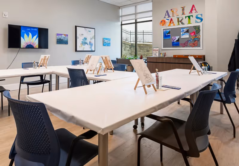 A bright art activity room with tables arranged in a U-shape, each table covered with white paper and equipped with small easels holding drawing sheets, paintbrushes, and colored pencils. The room has blue chairs around the tables, a large window letting in natural light, and a wall decorated with colorful letters spelling 'ARIA ARTS' along with artwork and butterflies on a bulletin board.