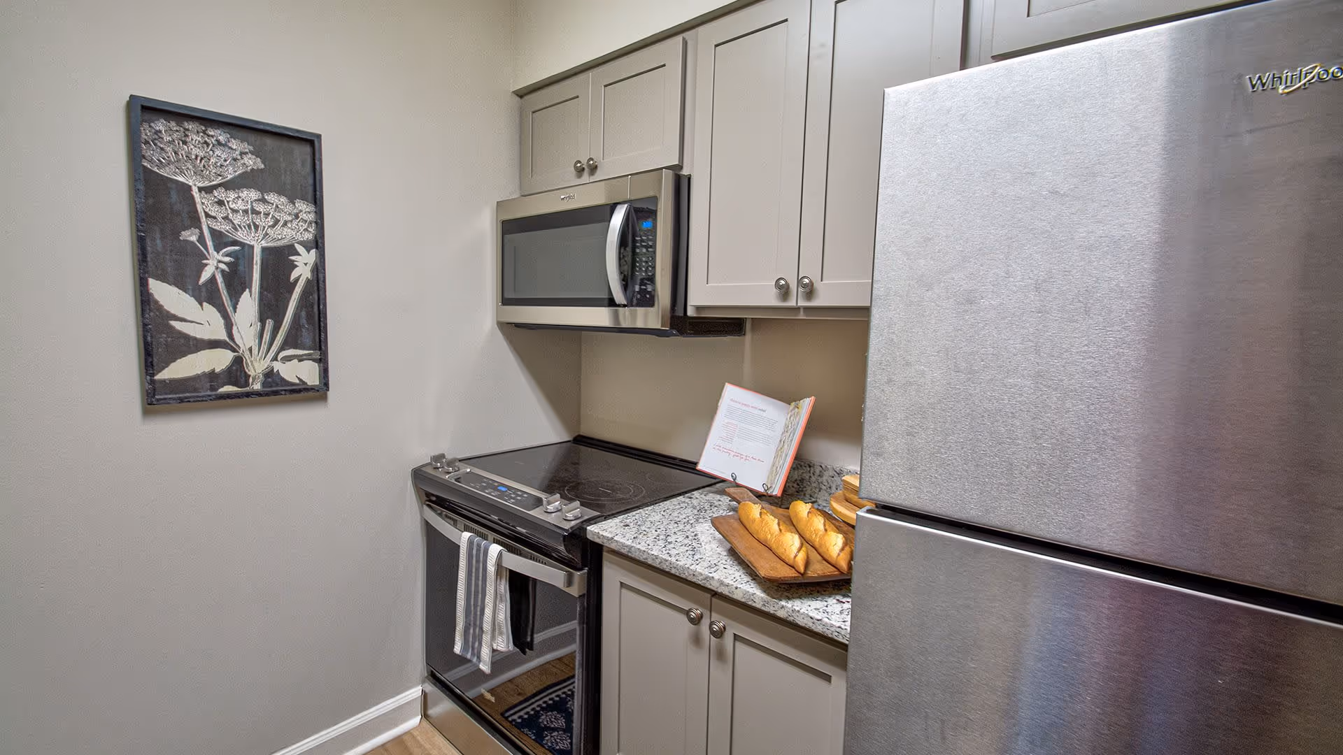 A small kitchen corner featuring a stainless steel Whirlpool refrigerator, an electric stove with a striped towel hanging on the handle, and a microwave oven mounted above the stove. There is a granite countertop with a wooden tray holding three baguettes and an open cookbook. A framed botanical artwork hangs on the adjacent wall.