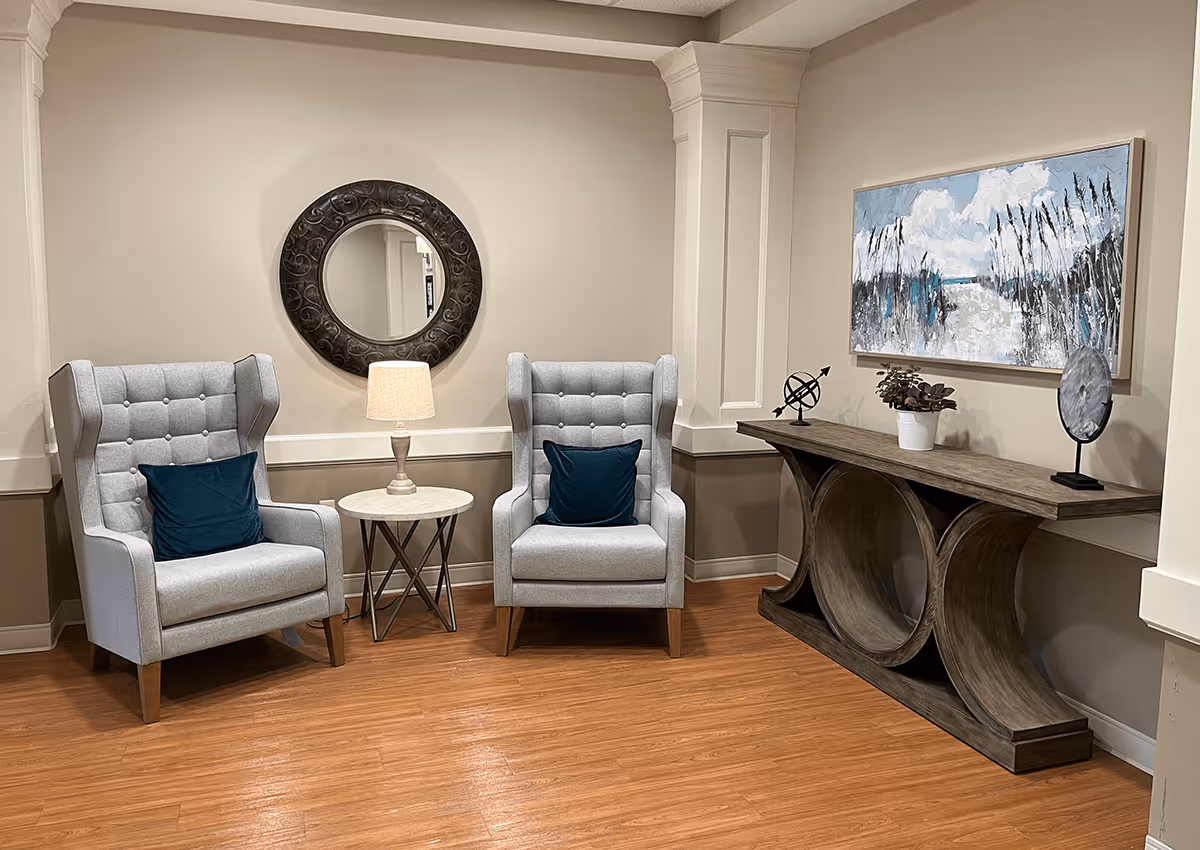 A cozy seating area with two gray upholstered armchairs, each with a dark blue cushion. Between the chairs is a small round table with a lamp on it. Behind the chairs is a round decorative mirror on the wall. To the right is a wooden console table with decorative items including a potted plant, a small sculpture, and a round mirror. Above the console table hangs a painting depicting a nature scene with clouds and tall grasses. The floor is wooden and the walls are painted beige with white trim.