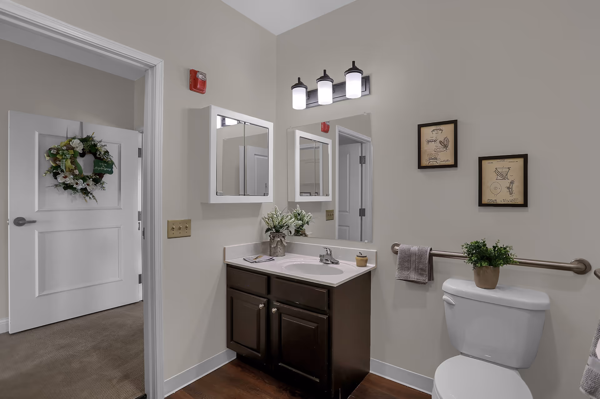 A clean and modern bathroom featuring a white toilet with a small potted plant on top, a dark wood vanity with a white countertop and sink, a wall-mounted mirror and medicine cabinet above the sink, and a towel hanging on a grab bar next to the toilet. The walls are light-colored with two framed pictures above the toilet. The bathroom door is open, showing a carpeted room with a white door decorated with a floral wreath.