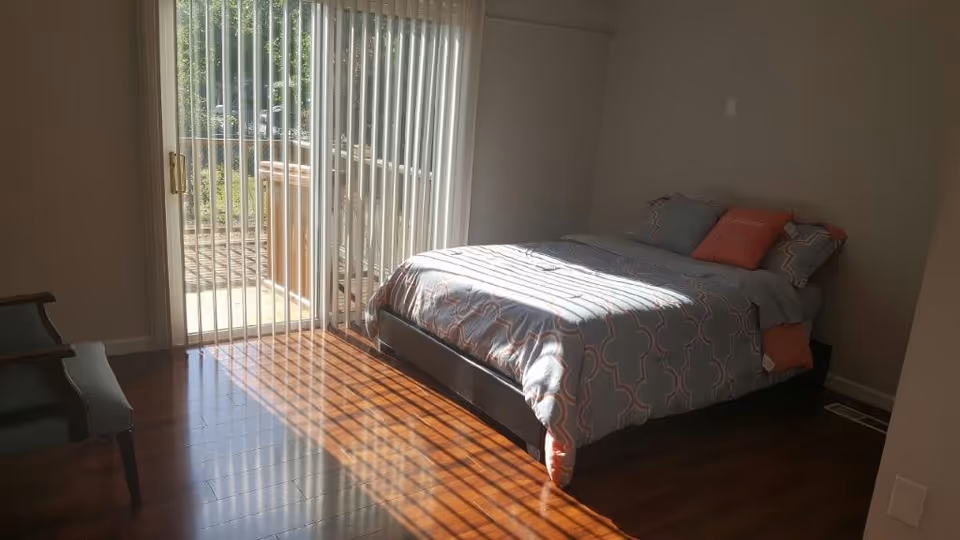 A bedroom with a bed covered in a gray and orange patterned comforter and pillows. Sunlight streams through vertical blinds covering a sliding glass door that leads to an outdoor area. There is a wooden chair with a cushion on the left side of the room and hardwood flooring throughout.