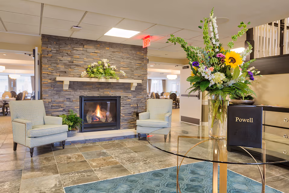 A cozy seating area in a senior living facility with two light blue armchairs positioned on either side of a stone fireplace with a fire burning. A glass table with a large floral arrangement and a book labeled 'Powell' is in the foreground. The background shows a dining area with tables and chairs.
