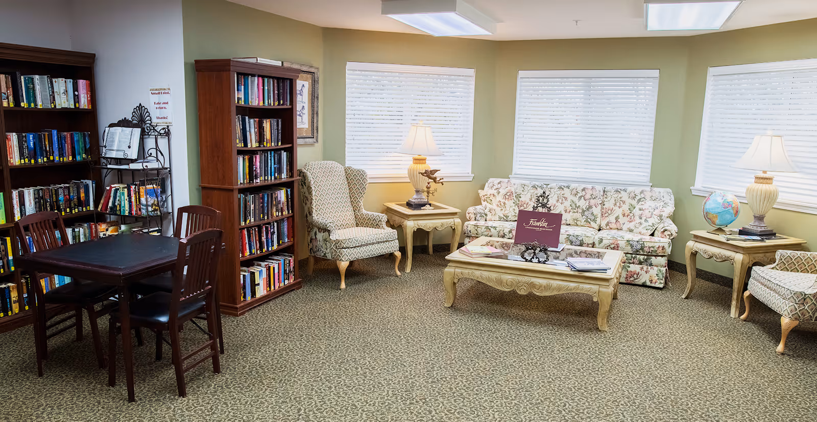 A cozy senior living community room with bookshelves filled with books, a small wooden table with four chairs, two patterned armchairs, a floral sofa, two side tables with lamps, a coffee table with books and a decorative item, and windows with white blinds.