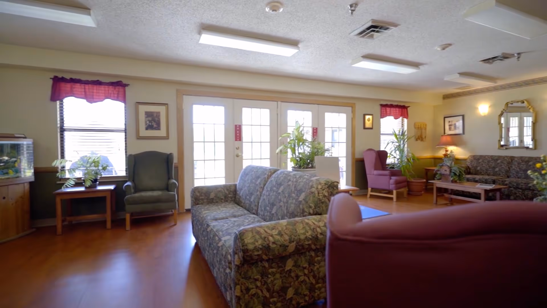 A well-lit living room area with floral patterned couches, armchairs, wooden tables, potted plants, and large windows with red valances allowing natural light to enter. The room has wooden flooring and soft yellow walls with framed pictures and a mirror.