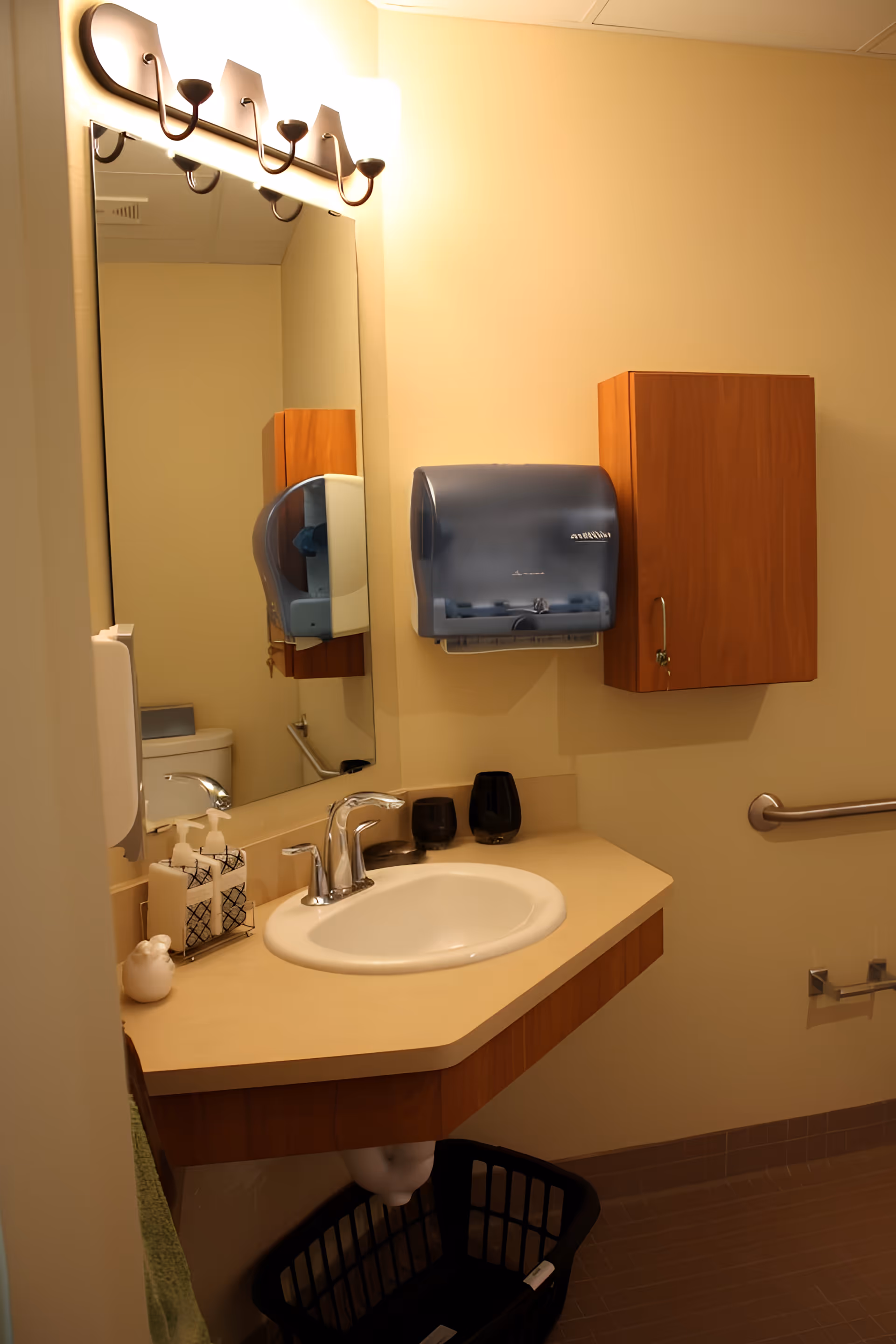 A clean bathroom with a beige countertop and a white sink. Above the sink is a large mirror with a light fixture featuring four lights. On the countertop are two soap dispensers and two black cups. A blue paper towel dispenser and a wooden cabinet are mounted on the wall. A grab bar is installed on the wall next to the sink, and a black laundry basket is placed underneath the sink.
