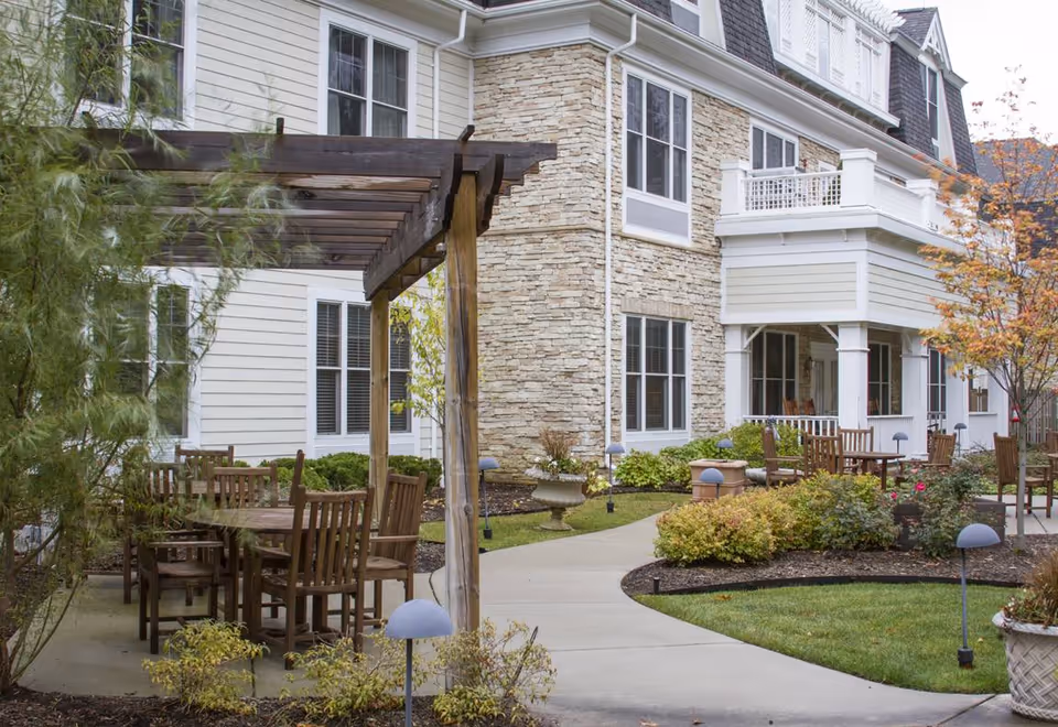 Outdoor patio area at Sunrise of Bloomfield Hills featuring wooden tables and chairs under a wooden pergola, surrounded by landscaped bushes and plants, with a multi-story building with stone and siding exterior in the background.