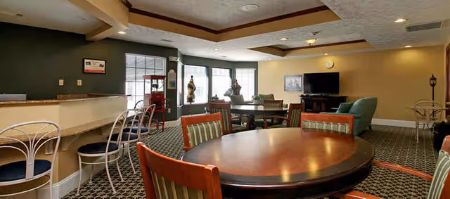 Communal dining and lounge area with round tables and chairs, a bar counter with stools, and a TV near bay windows.
