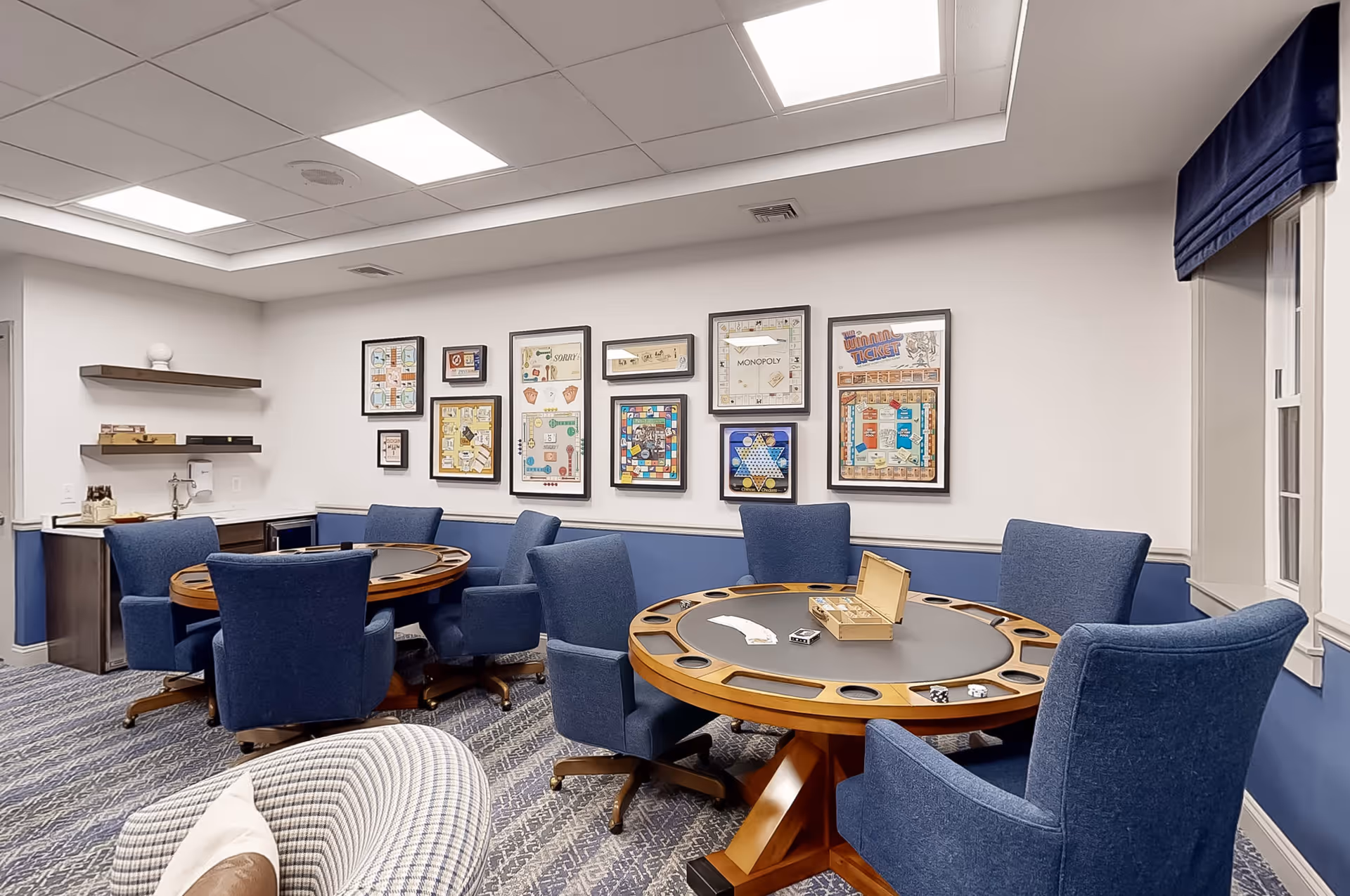 A game room with two round wooden tables designed for playing board games, surrounded by blue upholstered swivel chairs. The walls are decorated with framed vintage board game posters. There is a small counter with shelves and a sink in the corner, and a window with a blue valance on the right side.