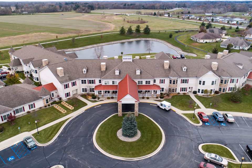 Aerial view of a senior living facility with a U-shaped building, central circular driveway and red-roofed entrance, parking lot, and a pond behind.