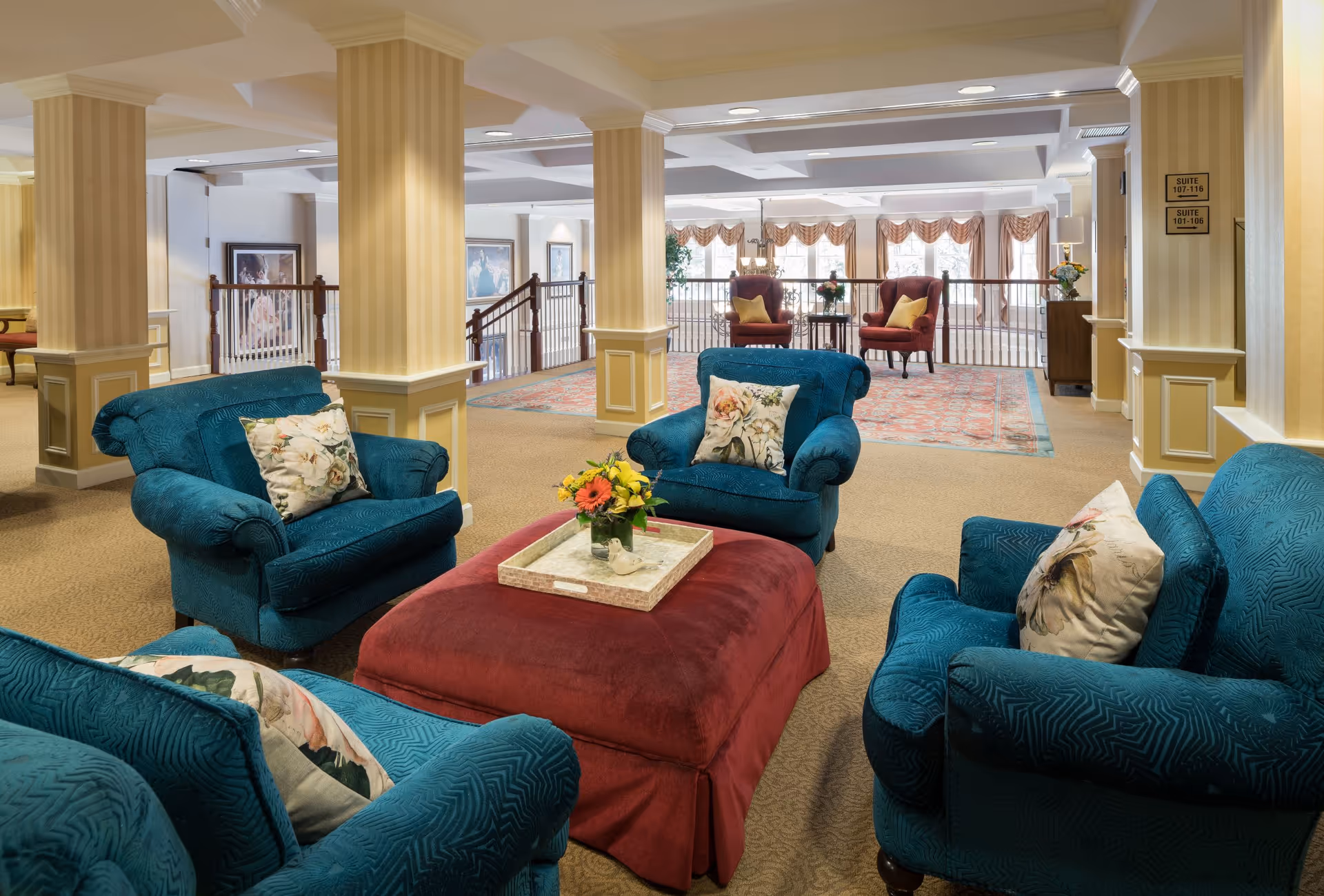 A cozy senior living common area with four teal upholstered armchairs arranged around a large red ottoman with a tray holding a small flower arrangement. The space features beige carpeting, cream-colored walls with decorative columns, and large windows with draped curtains letting in natural light. In the background, two burgundy armchairs with yellow pillows sit on a patterned rug near a railing overlooking a lower level.