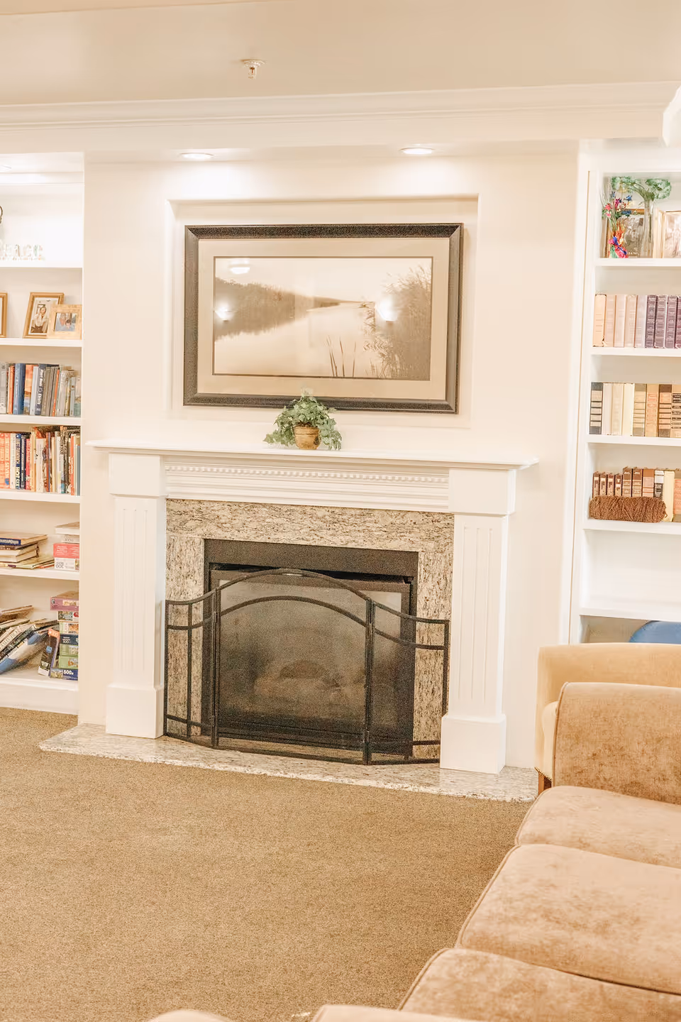 A cozy living room area featuring a white fireplace with a granite surround and a black metal screen. Above the fireplace hangs a framed landscape painting. On top of the mantel is a small potted plant. Built-in white bookshelves flank the fireplace, filled with books and decorative items. Part of a beige sofa is visible in the foreground.